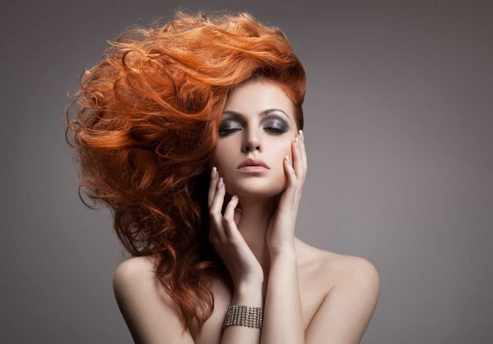 Red-haired woman with dramatic makeup posing with eyes closed and hands near face.